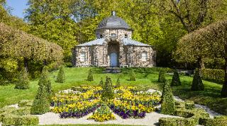 Ein barockes Lustschlösschen im Felsengarten Sanspareil. Im Vordergrund ist ein frühlingshaft bepflanztes Blumenparterre zu sehen. 