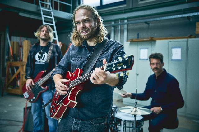 Ein Mann mit Gitarre schaut in die Kamera, im Hintergrund sitzt jemand am Schlagzeug und jemand mit Bass.