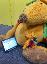 Ein großes Maskottchen und eine kleine Kuschelausgabe sind auf dem Boden. Vor ihnen ist ein Tablet.