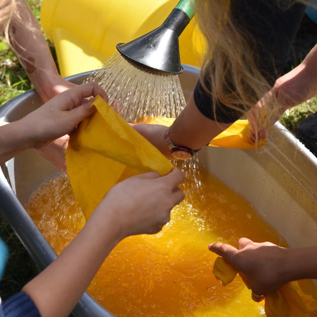 Gelbes Wasser ist in einer grauen großen Plastikwanne. Mehrere Kinder beugen sich über die Wanne. Ein Kin hält ein gelbes Stück Stoff zwischen den Händen. Mit einer Gießkanne wird Wasser in die Wanne gegossen.