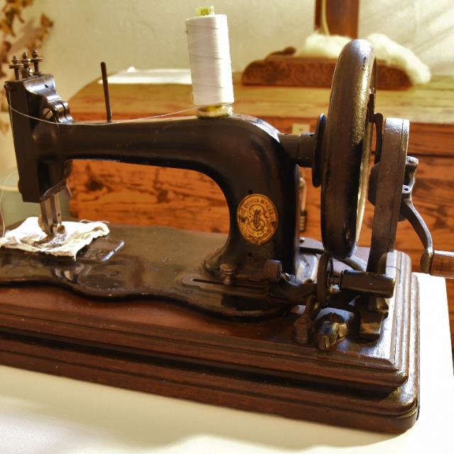 Auf einem hellen Tisch steht eine alte Nähmaschine. Eine weiße Rolle Nähgarn ist auf ihr befestigt. An der rechten Seite der Nähmaschine befindet sich eine Kurbel aus Metall.  