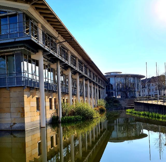 Ein Gebäude mit vielen Fenstern. Im Hintergrund ist der runde Sitzungssaal mit Glasfront. Vorne im Bild ist ein Teich. Das Gebäude spiegelt sich im Wasser. 