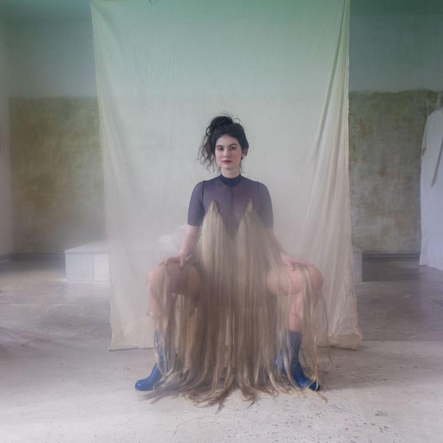 Eine dunkelhaarige junge Frau mit langem, beigen Rock sitzt breitbeinig vor einem blaugrünen Vorhang.