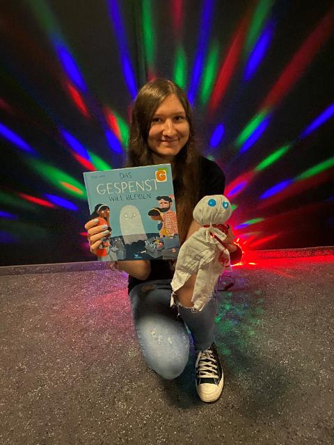 Eine junge Frau kniet hin und hält ein Bilderbuch und eine Geist-Puppe in der Hand. Hinter ihr sind bunte Lichter. 