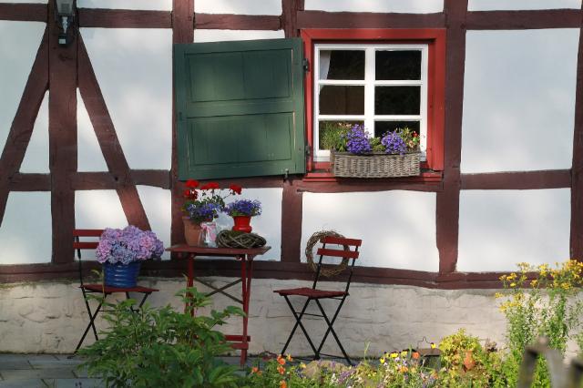 Ein Fachwerkhaus mit roten Fensterrahmen und grünen Fensterläden. Davor stehen Tisch und Stühle. Man sieht Blumen. 