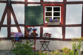 Ein Fachwerkhaus mit roten Fensterrahmen und grünen Fensterläden. Davor stehen Tisch und Stühle. Man sieht Blumen. 