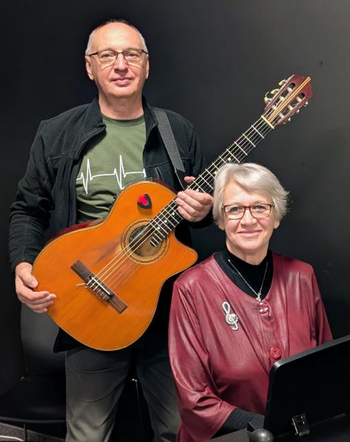 Ein älterer Mann mit einer Gitarre und eine Frau mit hellblonden Haaren, die vor ihm sitzt,.