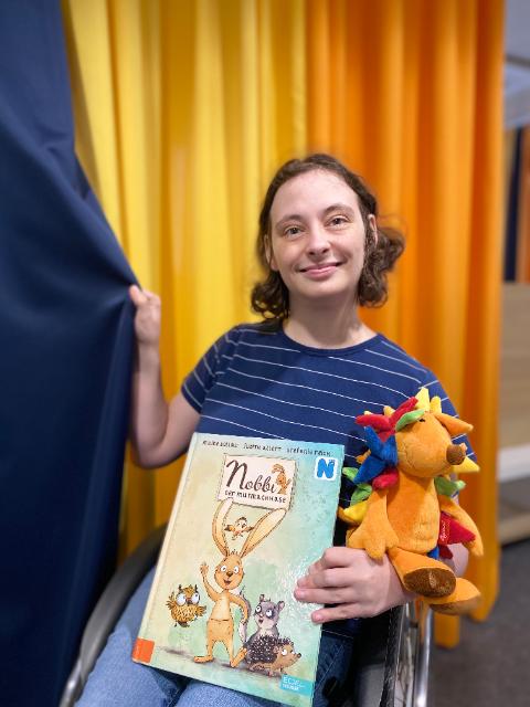 Junge Frau im Rollstuhl hält ein Buch und zieht an einem Vorhang