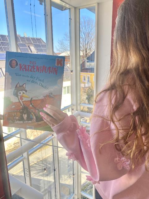 Eine junge Frau mit langen Haaren ist halb von hinten zu sehen, sie hält ein Bilderbuch in der Hand und steht vor einer Fensterfront.