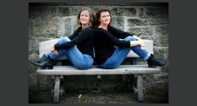 Anja und Alice sitzen Rücken an Rücken auf einer Bank