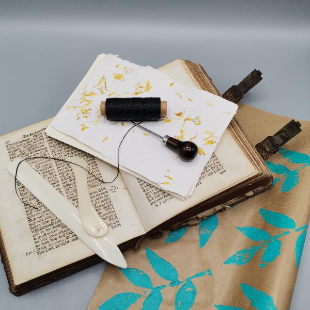 Auf einem hellen Untergrund liegt bunt bedrucktes Packpapier. Darauf liegt ein aufgeschlagenes Buch. Auf dem Buch liegt ein Papier mit getrocknetes Blüten, eine  Fadenrolle, eine spitze Ahle und zwei weiße Falzbeine. 
