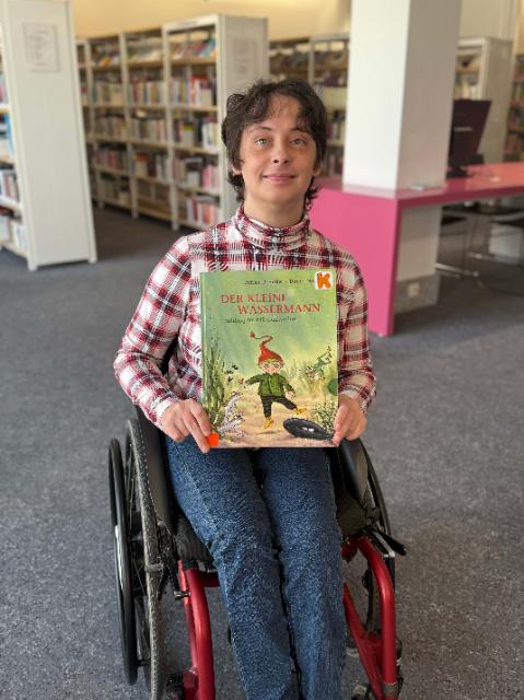Eine junge Frau im Rollstuhl und mit kurzen braunen Haaren, hält ein Bilderbuch in die Kamera.