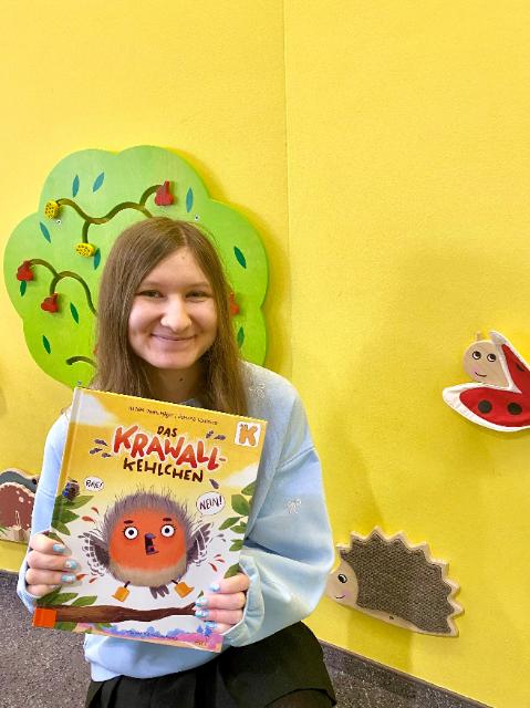 Eine junge Frau vor einer gelben Wand hält ein Bilderbuch in der Hand und lächelt.