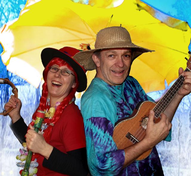 Eine Frau mit roter Parücke und ein Bann mit Hut und Gitarre lachen in die Kamera.