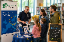 Ein Mann mit Brille an einem Infostand erzählt lächelnd einer Familie mit zwei Kindern etwas. 