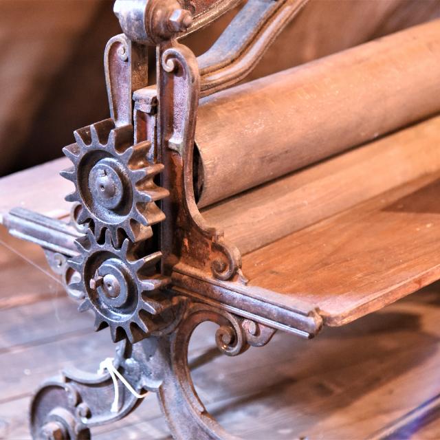 Ein Detailfoto zeigt zwei aufeinander liegende Walzen mit Zahnrädern an der Seite. Es handelt sich um eine Wäschemangel.