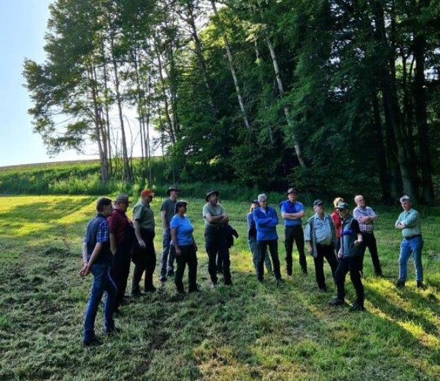 Eine Menschengruppe steht in der Wiese und hört einem Vortragenden zu.