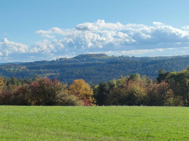 Der Betrachter steht auf einer grünen Wiese. Der Blick geht ins Weite, Himmel blau mit Wolken. Man sieht Wälder, teils herbstlich verfärbt. Im Hintergrund ein Tafelberg.
