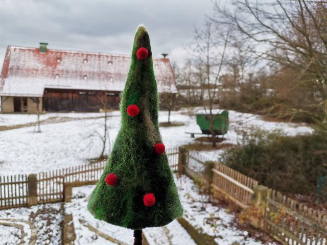 Ein grüner gefilzter Weihnachtsbaum ragt ins Bild. Rote Kugeln sind an ihm befestigt. Im Hintergrund ist eine Scheune und ein Garten. Überall liegt Schnee. 