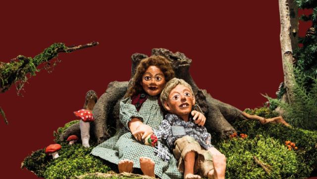 Die Marionetten Hänsel und Gretel sitzen im Wald vor einem Baumstamm 