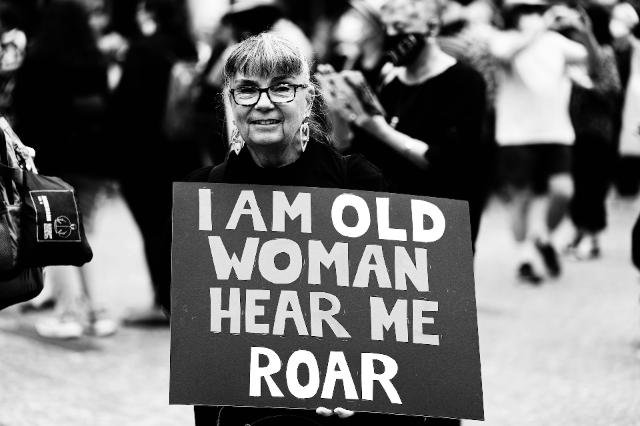 Ältere Frau mit Schild auf einer Demo. Symbol für Stärke, Selbstbewusstsein und Sichtbarkeit von Frauen in den Wechseljahren.