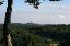 Mit Nadelbäumen bewaldete Hügellandschaft unter blau-weißem Himmel, im Hintergrund Blick auf einen Fernsehturm.