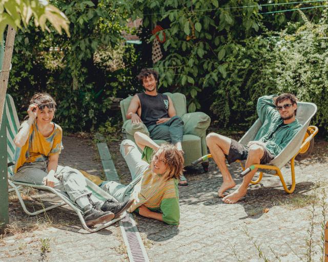 4 Leute in der Sonne im Grünen, 3 auf Gartenstühlen, eine liegt davor.