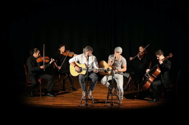 Graceland sitzt mit ihren Gitarren vor Violinspielern