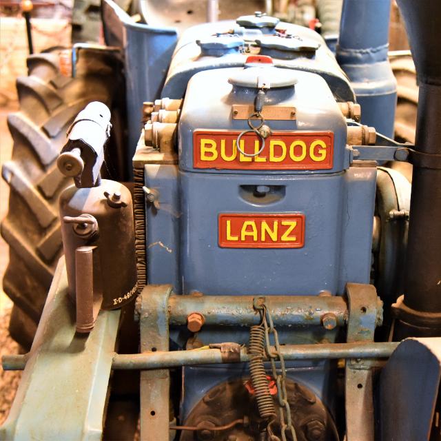 Ein blauer Traktor steht im Bildmittelpunkt. Er trägt eine große Aufschrift in Großbuchstaben auf seiner Schnauze. Die Aufschrift heißt: Lanz Bulldog. 