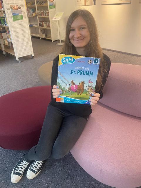 Eine junge Frau mit langen braunen Haaren sitzt auf einem Sofa und hält lächelnd ein Buch in die Kamera.