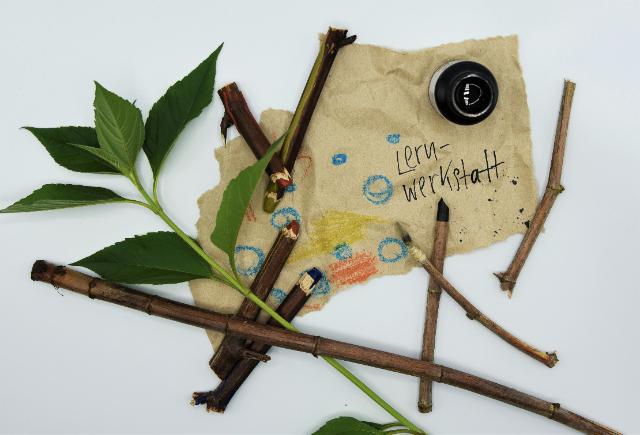 Auf einem hellen Untergrund liegt ein zerknittertes Stück Papier. Auf dem Papier steht "Lernwerkstatt" und daneben sind bunte Striche.  In einer Ecke des Papier steht ein offenes Tintenfass. Mehrere Stöckchen mit farbigen Spitzen liegen verstreut auf dem Untergrund. Der Stängel einer Pflanze mit grünen Blättern liegt neben dem Blatt.